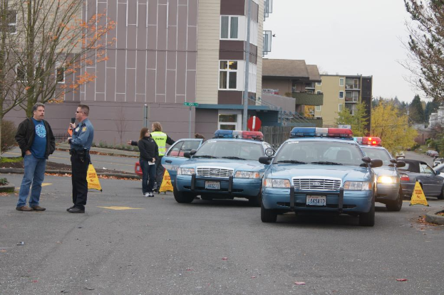 Autos auf einer Stra√üe mit vier Menschen in der N√§he, Geb√§ude mit Fenstern im Hintergrund, B√§ume und ein Warndreieck.