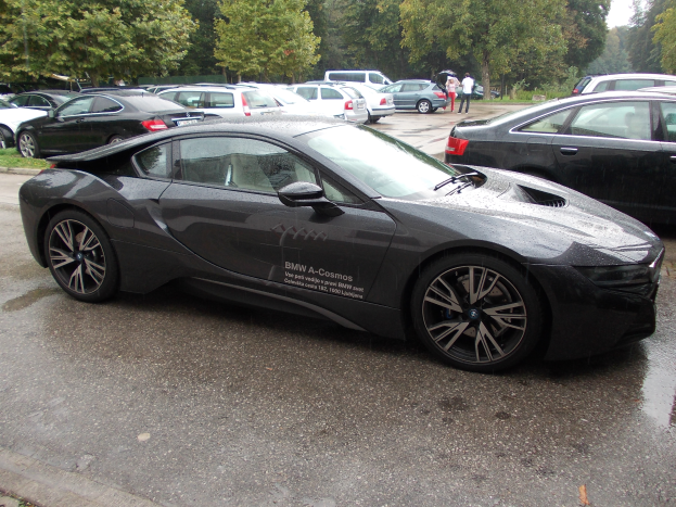 Ein schwarzer BMW i8 auf einem Parkplatz mit anderen Autos, Menschen in der Nähe, Bäumen und einem klaren blauen Himmel im Hintergrund.