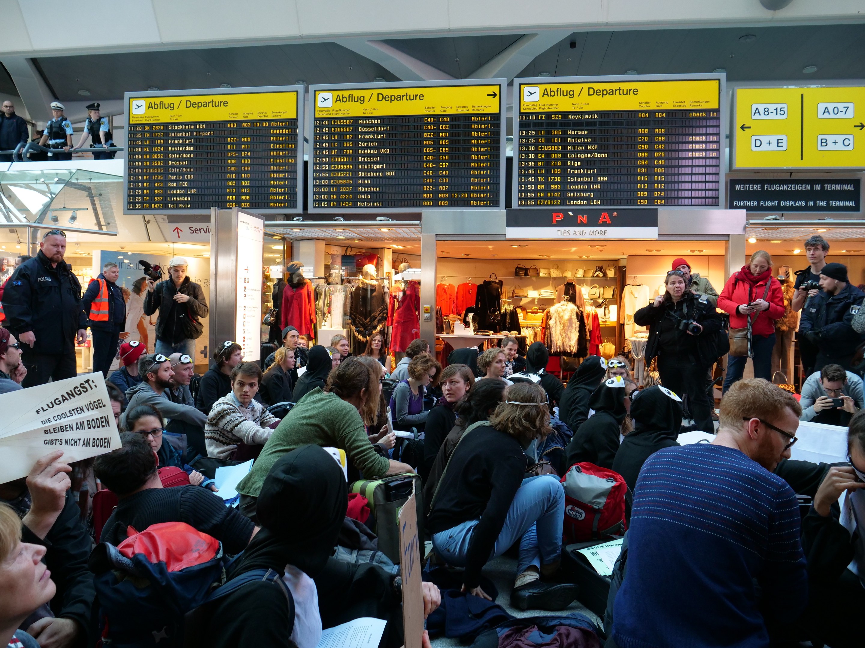 Eine große Gruppe von Menschen in einem Flughafen, einige sitzen mit Taschen und Papieren, andere stehen, mit Texttafeln und Schaufensterpuppen im Hintergrund, was auf eine Protestaktion hindeutet.