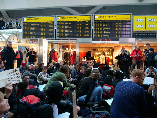 Eine große Gruppe von Menschen in einem Flughafen, einige sitzen mit Taschen und Papieren, andere stehen, mit Texttafeln und Schaufensterpuppen im Hintergrund, was auf eine Protestaktion hindeutet.