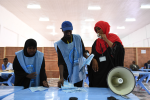 Eine Gruppe von Menschen steht um einen Tisch mit einem Megafon, Papieren und anderen Gegenständen herum, die scheinbar eine Wahllokal sind, mit anderen im Hintergrund unter beleuchteter Decke sitzend und stehend.