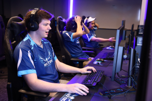 Gruppe junger Männer mit Headsets, die an Computern mit Monitoren, Tastaturen und Mäusen auf einem Schreibtisch spielen, mit beleuchteter Wand im Hintergrund.