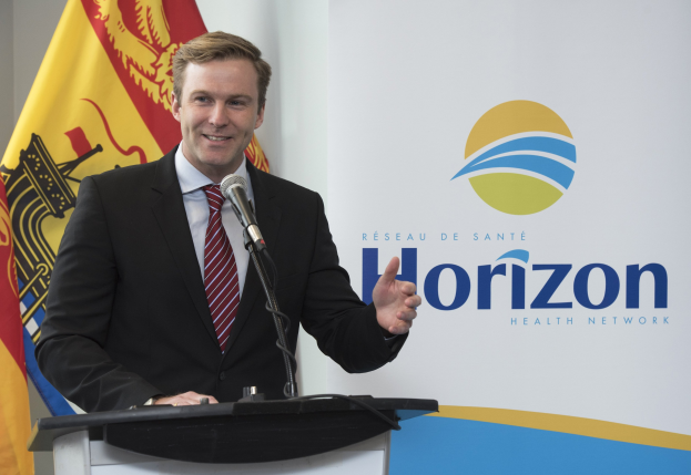 Ein Mann mit einem freundlichen Gesichtsausdruck in einem schwarzen Blazer, einem weißen Hemd und einer roten Krawatte steht an einem Rednerpult mit einem Mikrofon und einem Banner im Hintergrund.