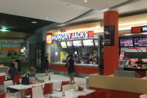 In einem Restaurant stehen Frauen an einer Essensbestellung auf einer Seite, während auf der anderen Seite Tische und Stühle mit einer Frau und einem Mann an einem der Tische angeordnet sind.