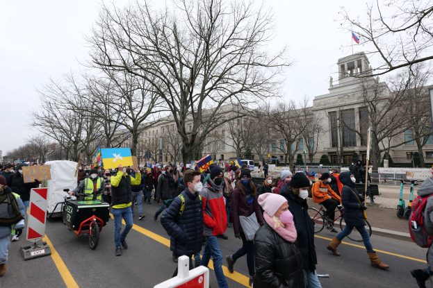Eine große Gruppe von Menschen marschiert auf einer Demonstration in Washington, D.C., mit Schildern und Transparenten, einige fahren Fahrräder, unter einem klaren blauen Himmel.