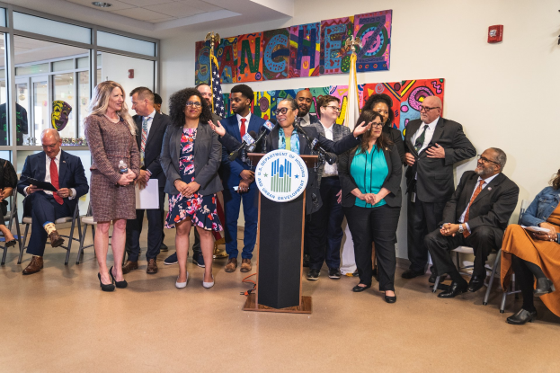 Eine Gruppe von Menschen steht vor einem Podium mit einem Mikrofon und einer Tafel mit dem Text "Sancho-Stadtrat tritt dem Stadtrat bei", während andere auf Stühlen sitzen oder in der Nähe stehen; Flaggen, ein Feuerlöscher und Deckenleuchten sind an der Rückwand zu sehen.