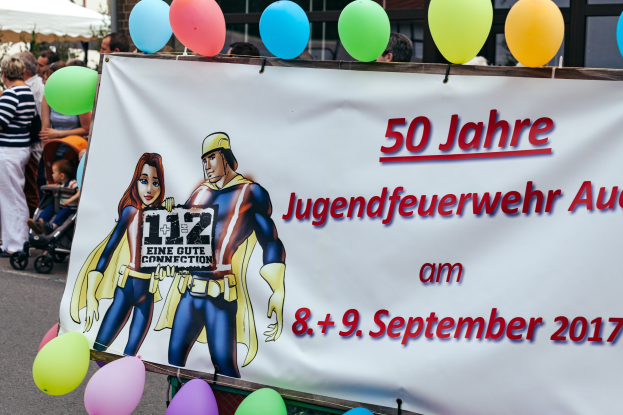 Eine Gruppe von Menschen steht auf der Straße und hält Kinderwagen, mit einem Banner "50 Jahre Jugendfeuerwehr August 2017", Ballons, ein Zelt, einen Zaun, Bäume und ein Gebäude im Hintergrund.