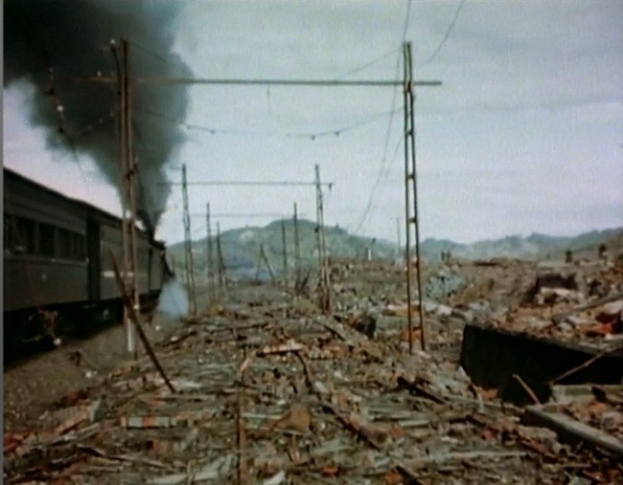 Ein Zug fährt durch ein geschädigt Gebiet mit Rauch, der aus ihm aufsteigt, umgeben von Strommästen und -drähten, Höhen und einem klaren Himmel im Hintergrund und verstreuten Trömmern am Boden.