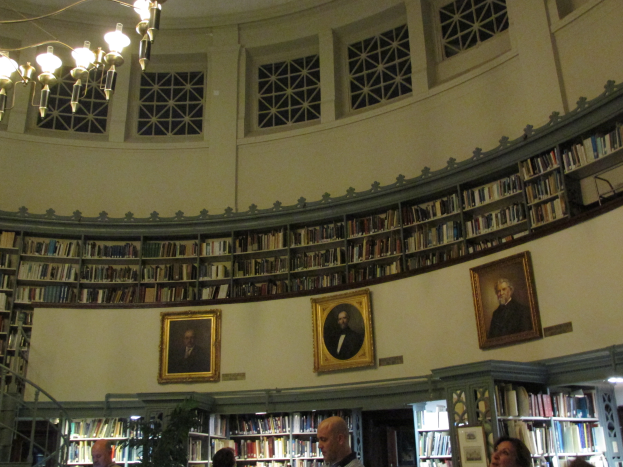 Ein großer Raum mit zahlreichen Bücherregalen, an der Wand hängenden Fotorahmen, einer Deckenleuchte, einer Eckpflanze und ein paar Menschen, die an eine Bibliothek erinnern.