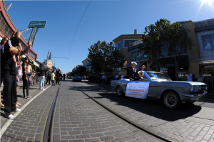 Ein Auto fährt eine Straße mit hohen Gebäuden entlang, mit Menschen auf den Gehwegen, einige halten Kameras, und Bäumen, Polen, Drähten und einem klaren blauen Himmel im Hintergrund.