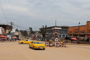 Ein gelbes Taxi fährt auf einer belebten Stadtstraße mit Gebäuden und Fenstern an den Seiten, während Fußgänger mit Schirmen auf dem Gehweg unter einem bewölkten Himmel gehen, mit Strommasten und Kabeln im Hintergrund.