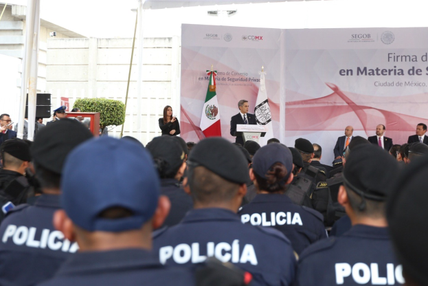 Eine Gruppe von Polizisten vor einer Menge mit Mützen, mit einer Person auf einem Podium in der Nähe einer Fahne und Flaggen, flankiert von einem anderen Podium, unter einem Himmel mit Pflanzen und Pfählen im Hintergrund.
