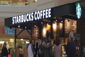 Eine Gruppe von Menschen steht vor einem Coffeeshop mit einem Namensschild, Logo, Menü und Lampen, mit einem anderen Geschäft daneben und einem Gang über dem Coffeeshop.