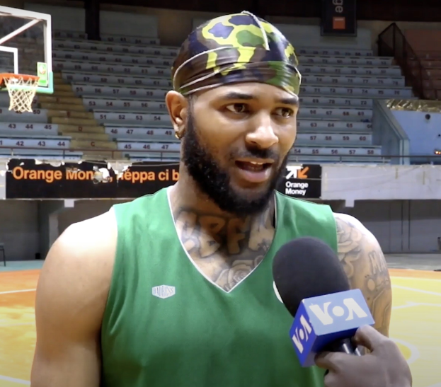 Ein Mann in einem grünen Tanktop, der in ein Mikrofon spricht, vor einem Basketballfeld mit einem Netz auf der linken Seite und Treppen im Hintergrund.