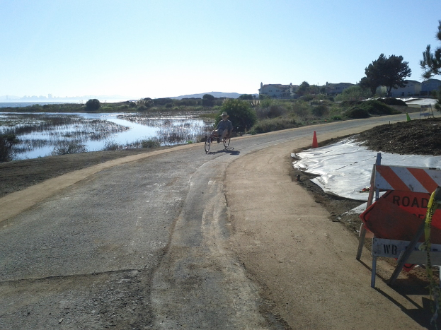 Ein Mann auf einem Fahrzeug auf einer Straße, mit einer Barriere und einem Verkehrskegel rechts und Wasser, Bäume, Gebäude und Himmel im Hintergrund.