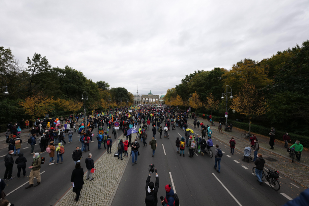Eine große Gruppe von Menschen marschiert auf einer von Bäumen gesäumten Straße in Berlin, hält Kameras während einer Demonstration, mit einem Gebäude und einem klaren Himmel im Hintergrund.