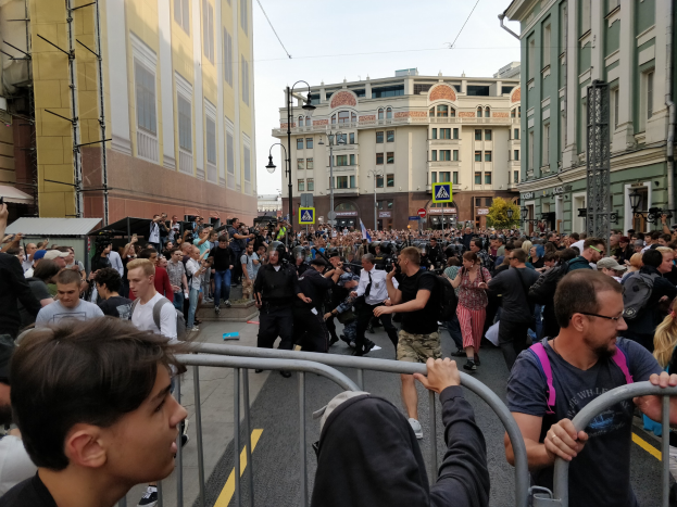 Eine große Menge steht hinter Metallabsperrungen auf einer von Gebäuden, Bäumen und Laternen gesäumten Stadtstraße, einige halten Kameras während einer Demonstration.