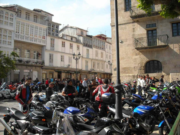 Ein cremefarbener Bau mit zahlreichen Fenstern, der an ein Backsteingebüude mit einer Metallbalustrade auf der rechten Seite angrenzt, mit mehreren parkenden Fahrrädern und Personen im Vordergrund, die sich unterhalten.