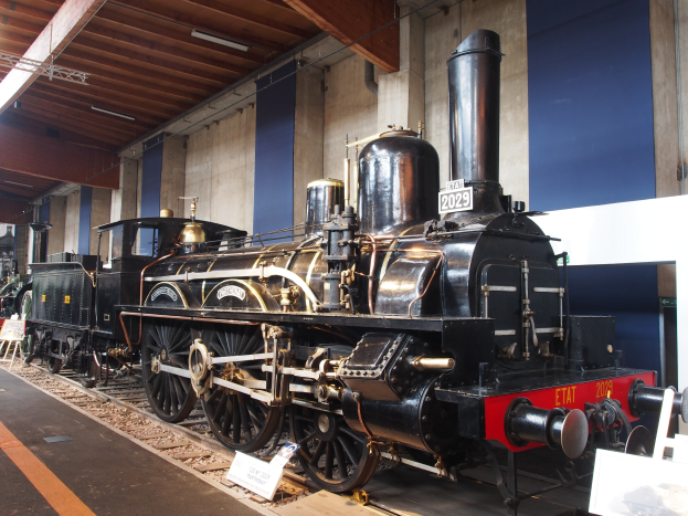 Alte Dampflokomotive in einem Museum ausgestellt, umgeben von Eisenbahnschienen, Informationsschildern und anderen Ausstellungsstücken, mit einer Wand und Deckenbeleuchtung im Hintergrund.
