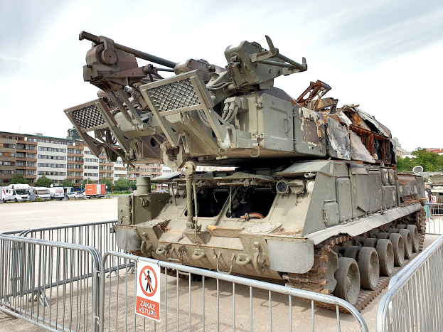 Ein gepanzerter Lkw steht vor einem Metallzaun mit einem daran befestigten Schild, mit Fahrzeugen, Bäumen, Gebäuden und einem klaren blauen Himmel im Hintergrund.