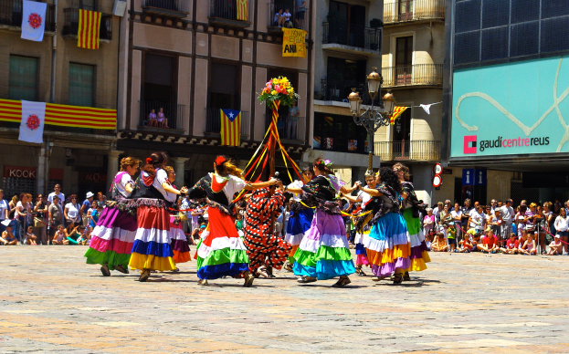 Eine Gruppe von Frauen in bunten Kleidern, die auf der Straße tanzen, umgeben von einer Menge Menschen, mit Gebäuden, Gelöndern, Balkonen, Flaggen, einer Laterne und einer Tafel mit Aufschrift im Hintergrund.