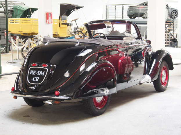 Schwarzes und rotes Auto in einer Garage mit anderen Fahrzeugen, Pfosten, Ketten und Gegenständen geparkt; Feuerlöscher an der Wand im Hintergrund.