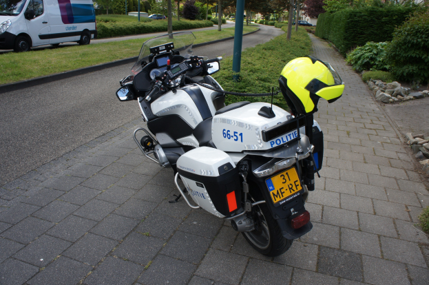 Ein auf der Seite der Straße geparktes Motorrad der Polizei mit einem Helm darauf, umgeben von Bäumen, Masten, Gebäuden, Pflanzen, Gras, Steinen und anderen Fahrzeugen.