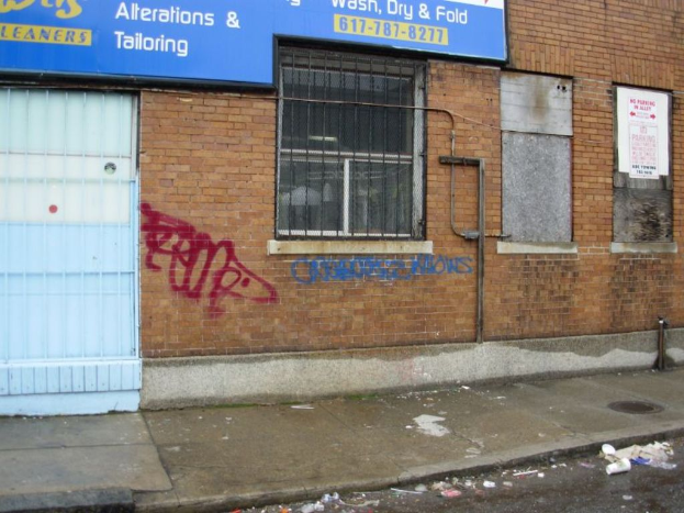 Eine Straßenszene mit einem Gehweg und einer Straße voller Müll, einer Backsteinmauer, Fenstern, Rohren, einem Tor und einer Werbetafel oben.