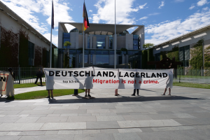 Eine Gruppe von Menschen hält ein Transparent mit der Aufschrift 'Deutschland, Lagerland Migration ist kein Verbrechen' vor einem Gebäude mit Fenstern, umgeben von Gras, einem Metallzaun, Pflanzen, Bäumen und Fahnen auf Stangen, unter einem bewölkten Himmel.