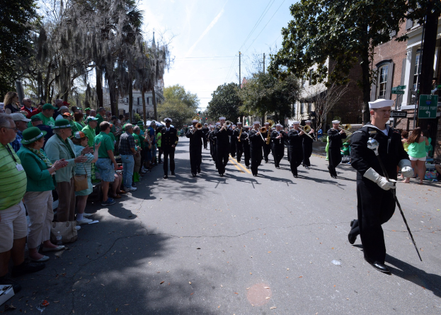 Eine Gruppe von Menschen in Mützen, einige mit Musikinstrumenten, marschiert auf einer Straße während eines St. Patrick's Day-Umzugs, mit Zuschauern auf der linken Seite und einem Gebäude, Bäumen und Strommasten im Hintergrund unter einem bewölkten Himmel.