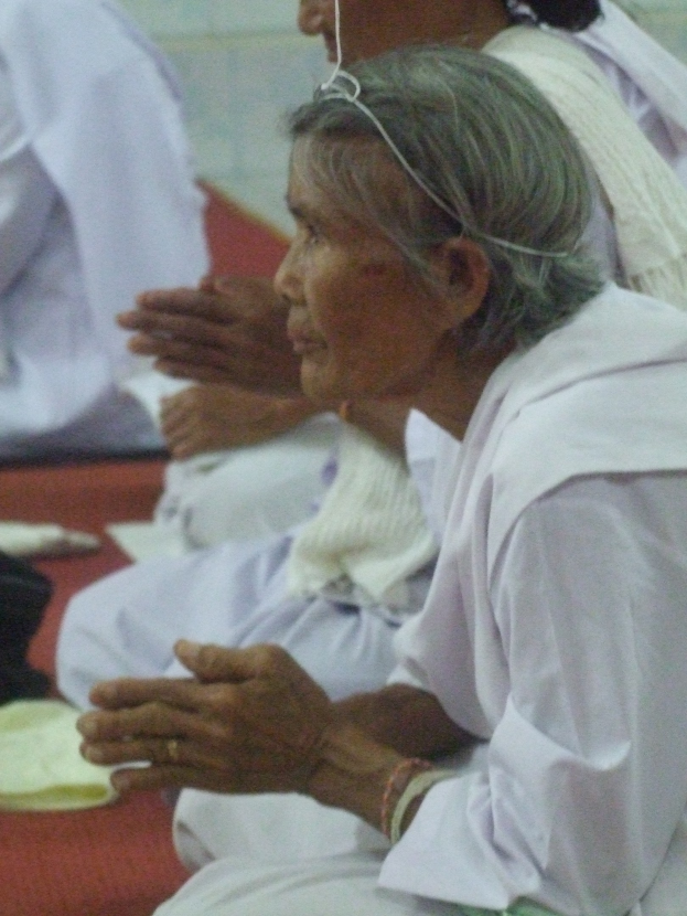 Alte Frau im Krankenhausbett liegend, Hände gefaltet, mit besorgtem Gesichtsausdruck und geschlossenen Augen, umgeben von Menschen im Hintergrund.