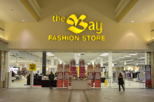 Ein Modegeschäft namens "bay fashion" in einem Einkaufszentrum mit Menschen, die gehen, Texttafeln an den Wänden, Säulen, Deckenleuchten und Hintergrundauslagen.