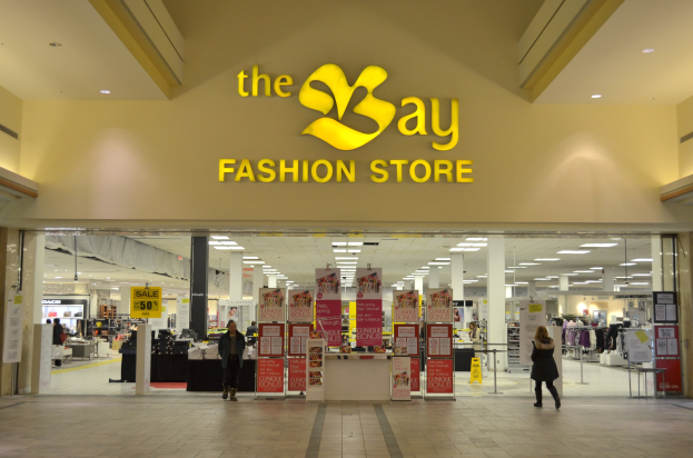 Ein Modegeschäft namens "bay fashion" in einem Einkaufszentrum mit Menschen, die gehen, Texttafeln an den Wänden, Säulen, Deckenleuchten und Hintergrundauslagen.