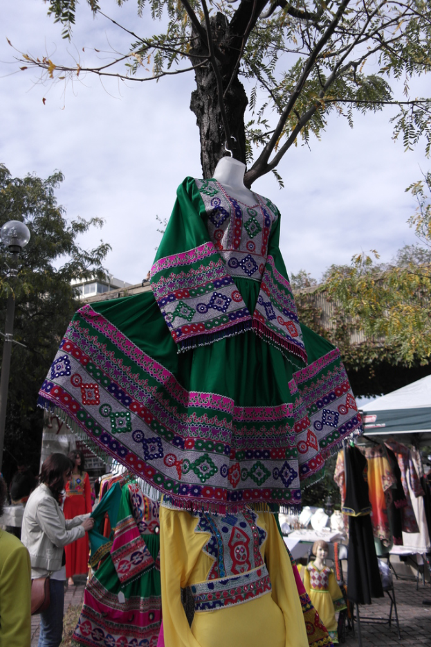 Eine Schaufensterpuppe steht inmitten eines belebten Einkaufsgebiets mit mehreren Geschäften, einer Person, die Kleider an einem Stab arrangiert, einem Gebäude und Bäumen im Hintergrund.