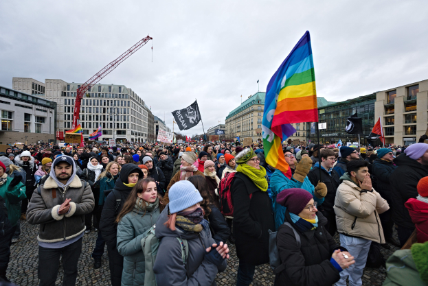 Große Gruppe von Menschen bei einer LGBTQ+-Rechte-Demonstration in Berlin, die Fahnen und Schilder schwenken, mit Gebäuden, einem Kran und einem bewölkten Himmel im Hintergrund.