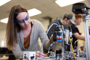 Eine Frau mit Schutzbrille arbeitet an einem Roboter in einem Labor, umgeben von anderen Personen und Gegenständen auf dem Tisch sowie Deckenbeleuchtung.