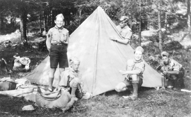 Schwarz-weißes Bild von Pfadfindern in Uniform, die sich um ein Zelt in einem bewaldeten Gebiet versammelt haben, einige sitzen auf dem Boden, während andere stehen, mit verstreuten Taschen und Ausrüstung.