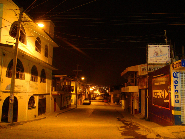 Eine Straßenszene mit Gebäuden und Strommasten auf beiden Seiten, parkenden Fahrzeugen vor den Gebäuden und einer Straße in der Mitte.