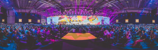 Eine große Menschenmenge sitzt auf Stühlen vor einer hell erleuchteten Bühne mit mehreren stehenden Personen, wahrscheinlich auf einem Online-Marketing-Rockstars-Event, umgeben von Bildschirmen.