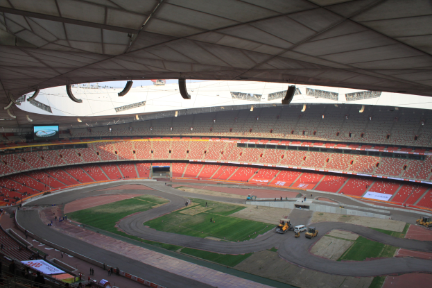 Großes Stadion voller Menschen, Fahrzeuge auf der Straße, Gras, Texttafeln, Geländer, ein Bildschirm und Deckenleuchten.