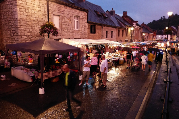 Eine Gruppe von Menschen, die auf der Straße gehen, einige mit Schirmen, mit Läden, die Spielzeug, Taschen und Gebäude mit Fenstern im Hintergrund zeigen, sowie Bäume, Straßenlaternen und Absperrungen an der Seite.