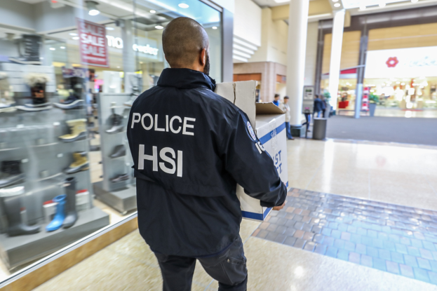 Ein Polizist in einer schwarzen Jacke trägt eine Kiste und geht durch ein Einkaufszentrum mit Menschen, einem Müllcontainer, Säulen und Deckenlampen im Hintergrund, während auf der linken Seite Schuhregale zu sehen sind.