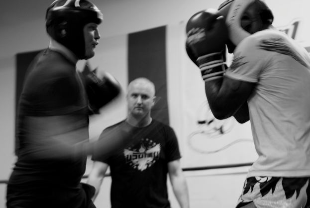 Zwei Boxer mit Helmen und Handschuhen kämpfen im Ring, mit einem Schiedsrichter zwischen ihnen und einem Banner im Hintergrund.