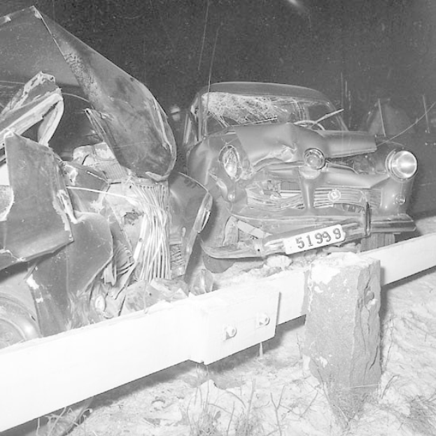 Schwarz-weiß-Foto eines demolierten Autos mit eingedellter Motorhaube und zersplitterten Fenstern, das auf Gras neben einem Zaun mit dunklem Hintergrund geparkt ist.