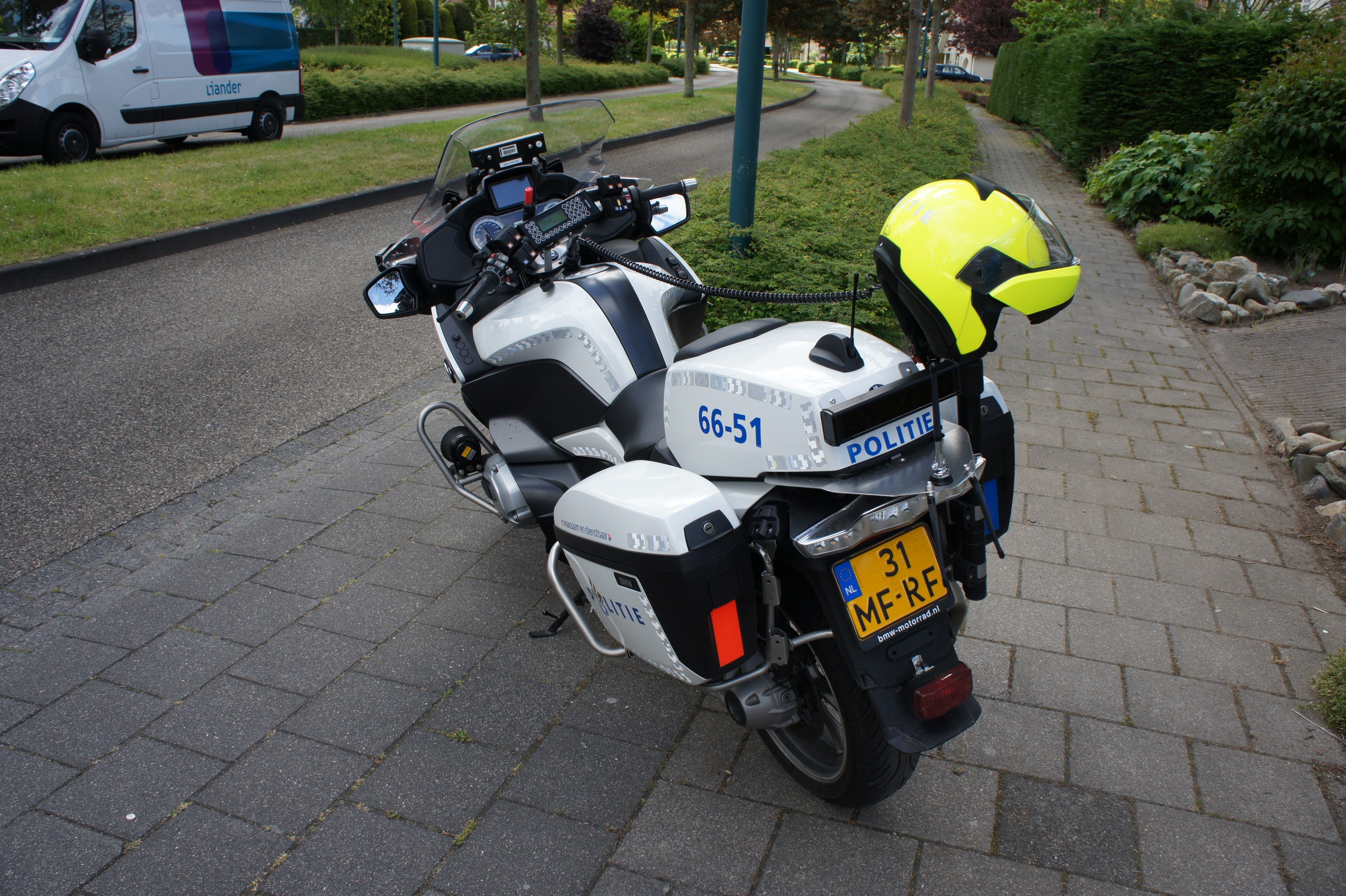 Ein Polizeimotorrad am Straßenrand geparkt mit einem Helm darauf, umgeben von Bäumen, Pfählen, Gebäuden, Pflanzen, Gras, Steinen und anderen Fahrzeugen im Hintergrund.