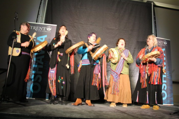 Eine Gruppe von Frauen in bunten Kleidern, die singen und Trommeln halten, mit einem schwarzen Vorhang und zwei blauen Roll-up-Plakaten im Hintergrund.