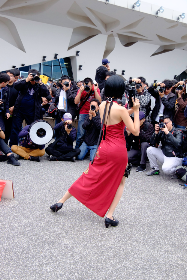 Eine Frau in einem roten Kleid tanzt vor einer Menge, einige halten Kameras und einer ein Megafon, mit einem beleuchteten Gebäude im Hintergrund.