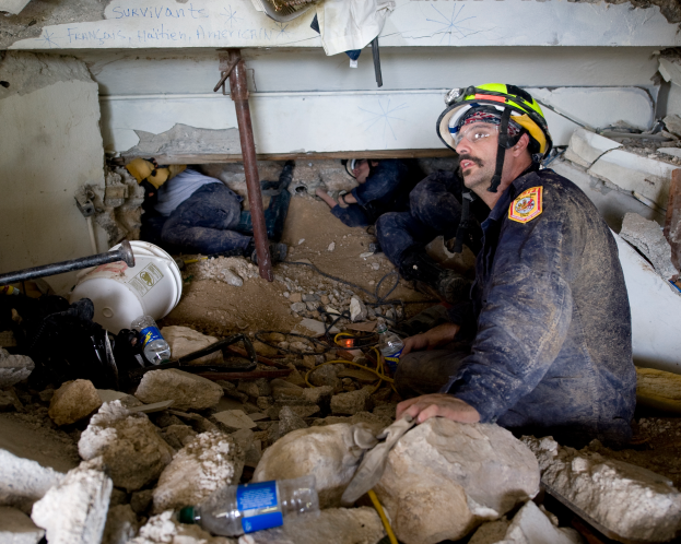 Ein Feuerwehrmann in einem Helm sitzt zwischen Trümmern, einschließlich Steinen und Flaschen, mit einer Wand im Hintergrund, auf der ein Text steht.