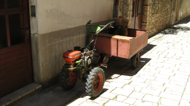 Ein Traktor mit einem Anhänger auf einer Straße, neben einem Haus mit sichtbaren Türen.