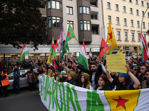 Eine große Gruppe von Menschen, die eine Straße entlanggehen und Fahnen und Plakate schwenken, mit einem Auto am Straßenrand, einem Baum links und Gebäuden mit Fenstern und Namensschildern im Hintergrund, was auf eine algerische Demonstration hindeutet.